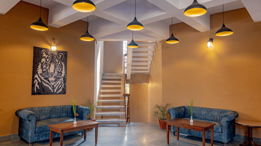 Indoor lounge at Estherea Bagh, Ranthambore, with upholstered sofas, wooden coffee tables, hanging pendant lights, and a staircase leading upstairs.