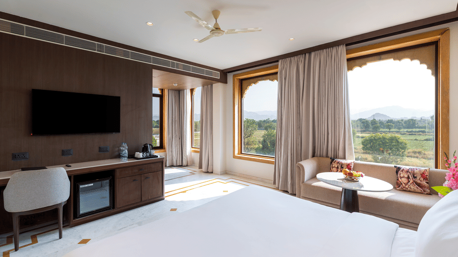 Room interior at EsthereaRaj Leela, Ranakpur, featuring a bed, a television on a wood-panelled wall, a white chair, and a large window with light-coloured drapes.