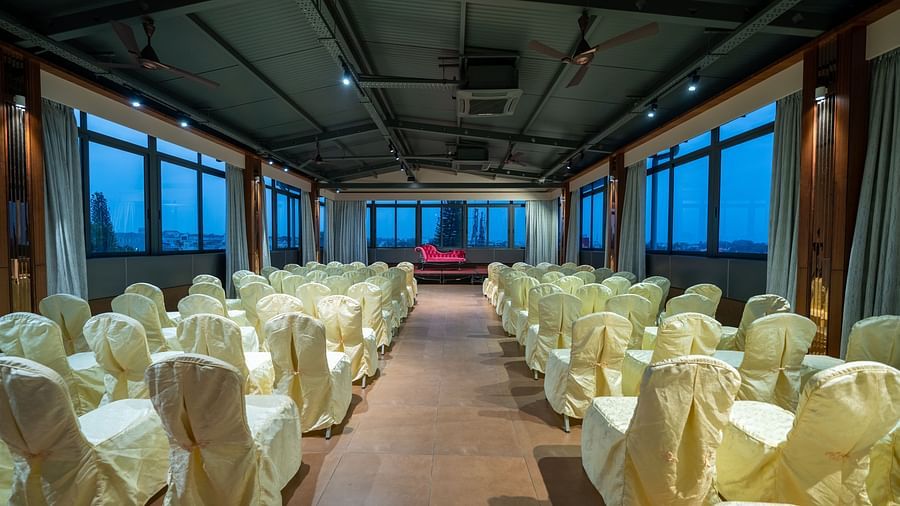 A view of the banquet hall at Hari Govinda's Express, Vijayanagar, with chairs arranged on either side of a central aisle leading to the stage, which features a cushioned sofa, while the curtains are drawn to the sides.