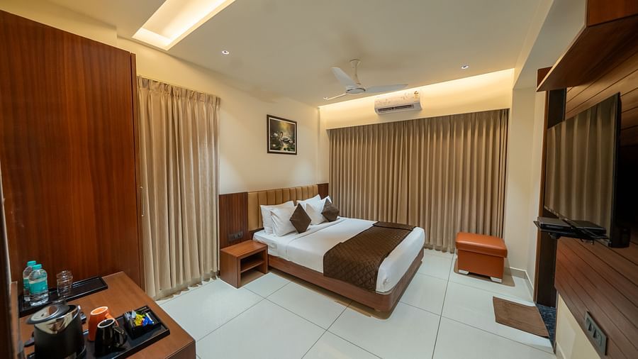 A spacious room at Hari Govinda's Express, Vijayanagar, featuring a large bed with a wooden headboard, a bedside table, a wall-mounted TV, and a wooden table with essentials, with curtains fully drawn to the side.