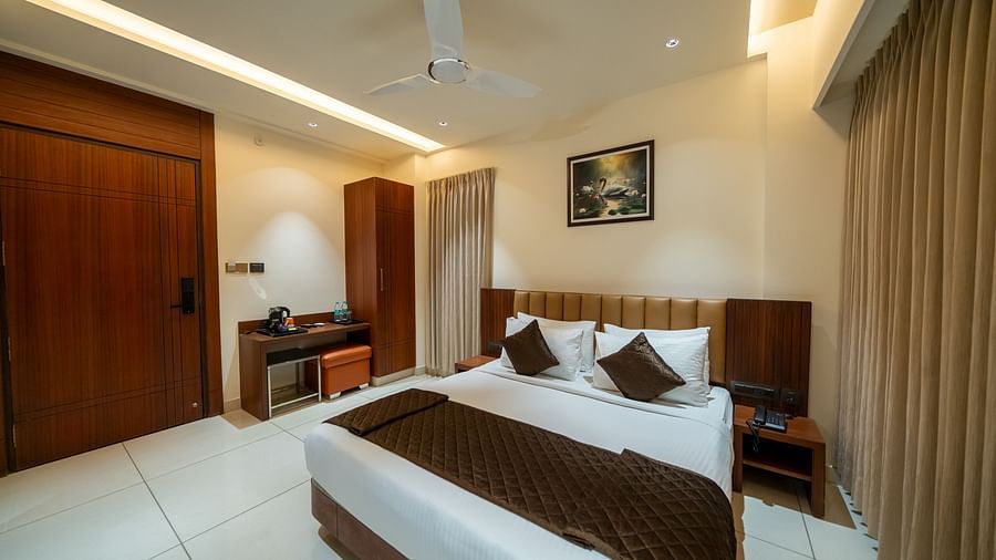Hari Govinda's Express, Vijayanagar, features a neatly made bed with a photo frame above, a table and cupboard to the left, and drawn curtains to the right.