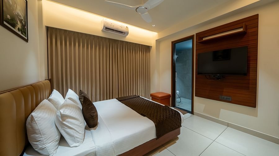 A side view of the bed at Hari Govinda's Express, Vijayanagar, with a photo frame above the headboard, fully drawn curtains, a doorway, and a wall-mounted TV beside it.