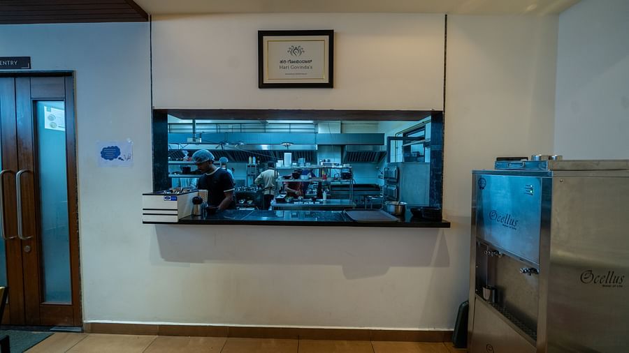 A rectangular glass on the wall at Hari Govinda's offering a view of the other side, with a photo frame above it and a metal water purifier beside it