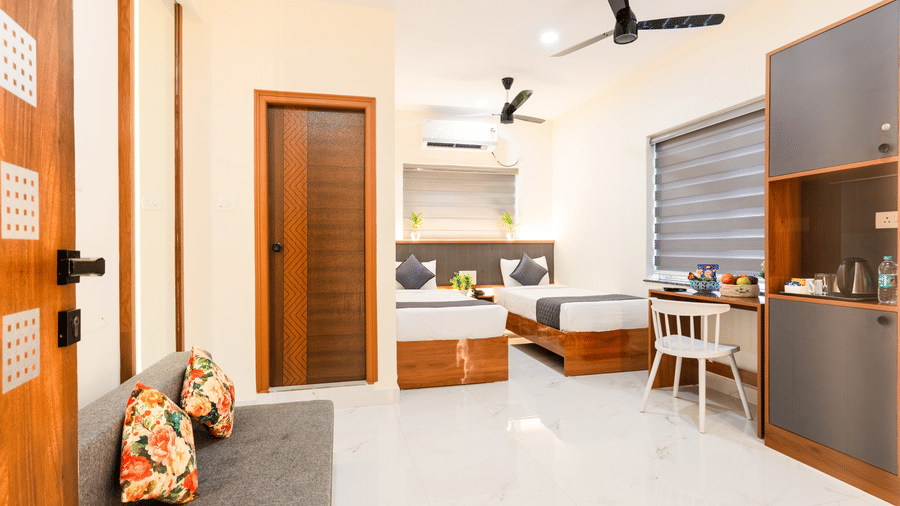 A room at Hotel Aurora Bliss, Gachibowli, featuring twin beds, spreads, pillows, a sofa, a fan, and a kitchenette.