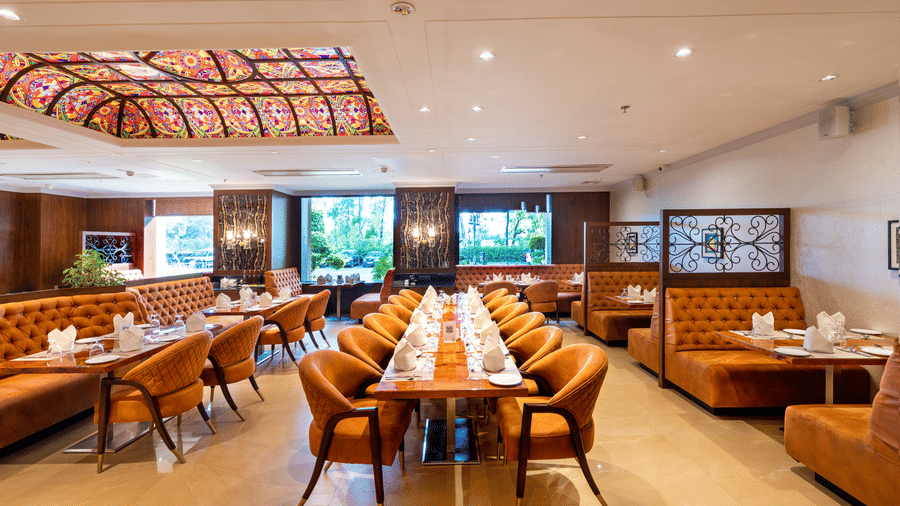 Restaurant dining hall featuring orange booths, wooden tables set with orange cloths, and a decorative ceiling panel at Hotel Hukam's Lalit Mahal.