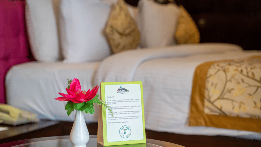 A neatly made bed with white linens, decorative pillows, and a bedside table holding a flower vase and a service card at Hotel Udai Median, our hotel in Udaipur city