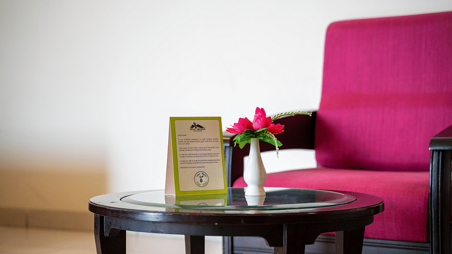 A small round table with a flower vase and a hotel service card, placed next to a armchair and a bed - Hotel Udai Median, Udaipur.