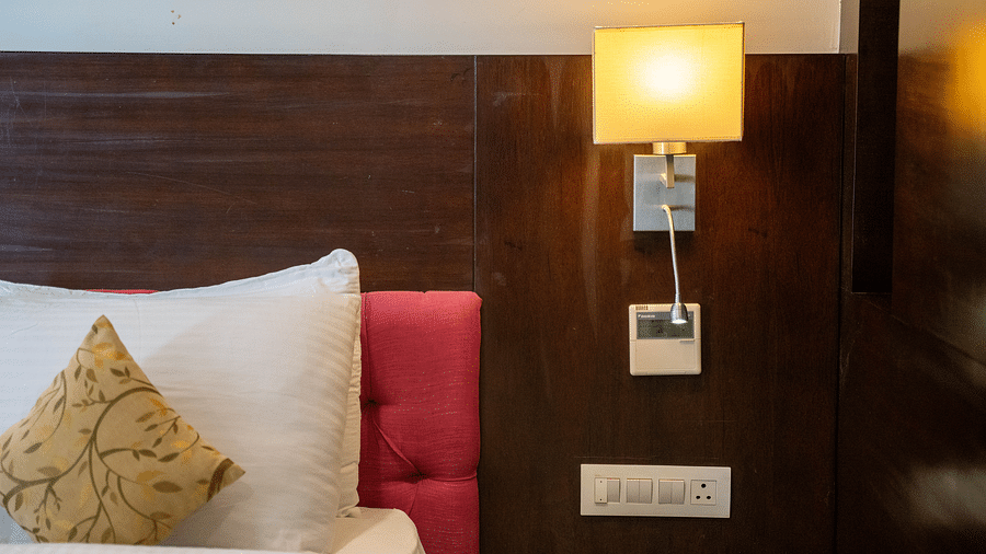 A cosy hotel bed with white linens, decorative pillows, and a bedside lamp on a wooden panel - Hotel Udai Median, Udaipur.
