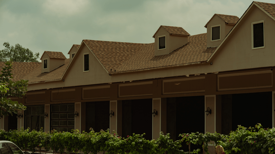 A tan building features multiple small dormer windows on a brown tiled roof, with a row of green potted plants lining the walkway below at Jivana Wellness Retreat, Bhopal.