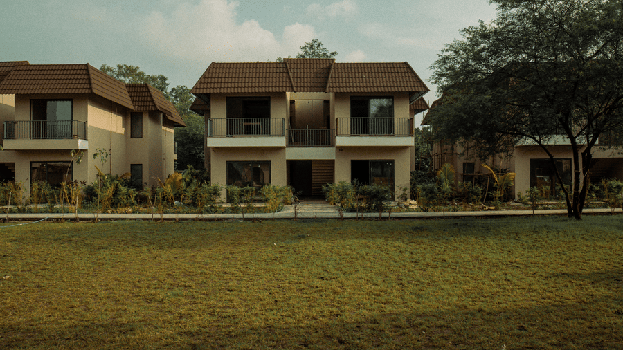 2 identical storey tan buildings with dark roofs stand behind a flat green lawn, separated by a central walkway and trees at Jivana Wellness Retreat, Bhopal.