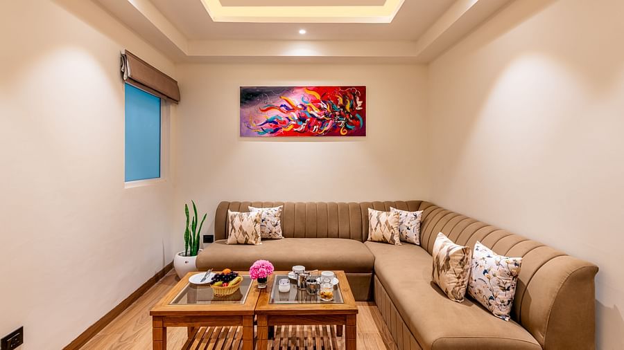 A living room with a sectional couch, a coffee table, and an abstract painting on the wall. It features a distinctive tray ceiling with lighting and a small window - Lords Eco Inn Dehradun.