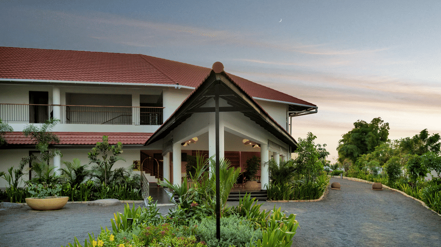 View of the building entrance at Mango Hill, Velankanni with a driveway and plants, and trees in the background.