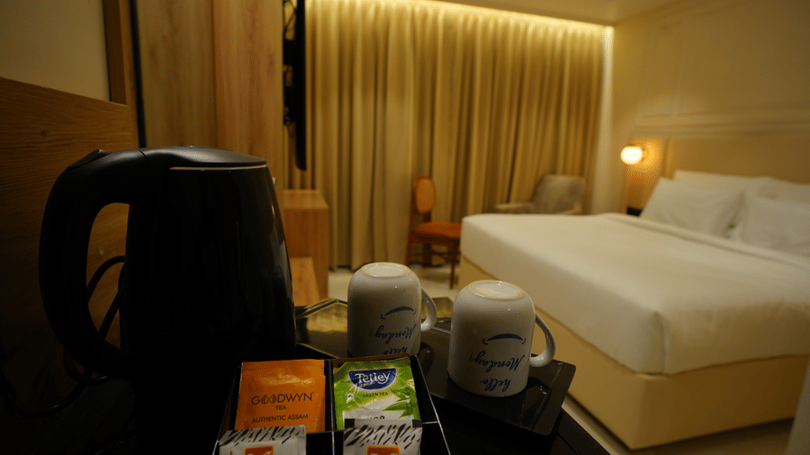  Kettle and mugs for room amenities at Monday Premium, Madhapur, with the double bed and gold-draped window blurred in the background.
