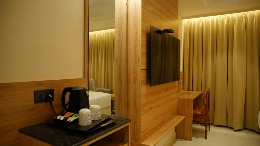  Modern guest room setup at Monday Premium, Madhapur, showing a TV on a wood-panelled wall and tea-making facilities nearby.