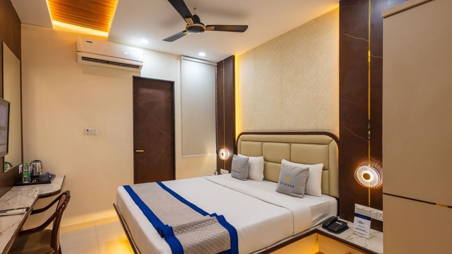 A room with a double bed, warm lighting, blue & beige runner, bedside lamps & a chair facing the wall at Pravasa Stay Dewas Naka, Indore.
