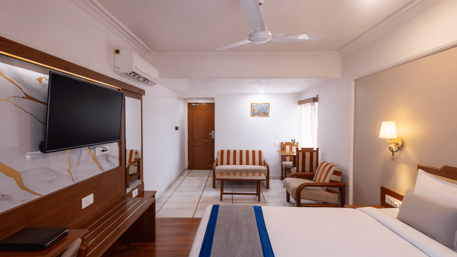A warm, spacious hotel room with a wooden floor, a large bed, and a seating area visible near the window at Pravasa Stay, Vadodara.