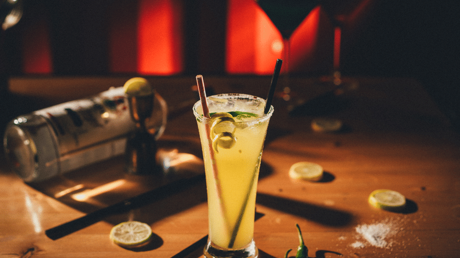 A tall glass of a yellow cocktail garnished with citrus slices sits on a dimly lit wooden bar next to a metal cocktail shaker at S Hotels, Chennai.
