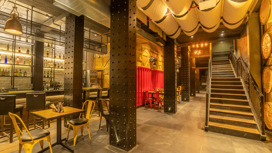 The interior of a dimly lit bar shows tables, chairs, and a staircase leading to an upper level at S Hotels, Chennai.