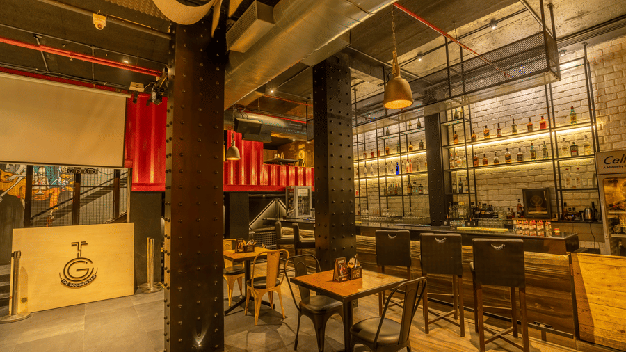 A dimly lit bar interior features wooden tables and tall stools in front of a counter with bottles of spirits displayed at S Hotels, Chennai.