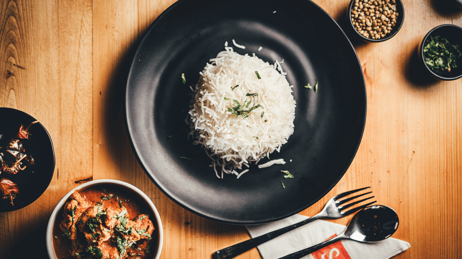 A plate of white rice garnished with black peppercorns sits next to small condiment bowls and cutlery on a wooden surface at S Hotels, Chennai.