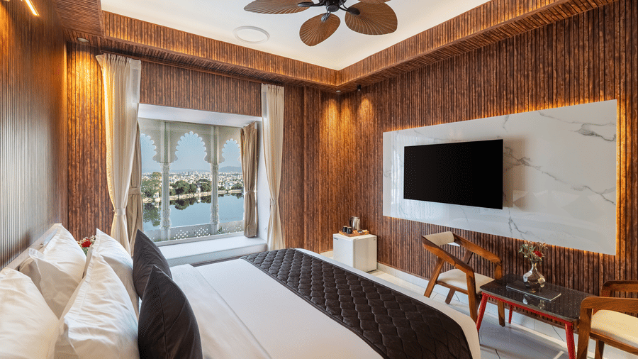 Modern hotel bedroom with a comfortable bed, a large window offering a lake view, and a wall-mounted TV on a marble panel.