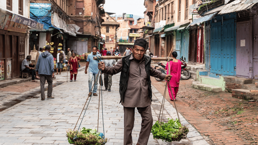 A local vendor carrying fresh green produce using a traditional wooden shoulder pole down a paved street in Bhaktapur, Nepal.