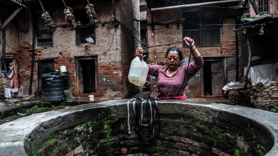 A local woman drawing water with a bucket from a communal stone well in a historic residential courtyard in Patan, Nepal.
