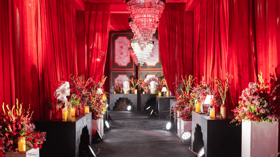An indoor wedding aisle is adorned with red fabric, flowers, and candles | The Qualia Resort Club & Brewery, Udaipur