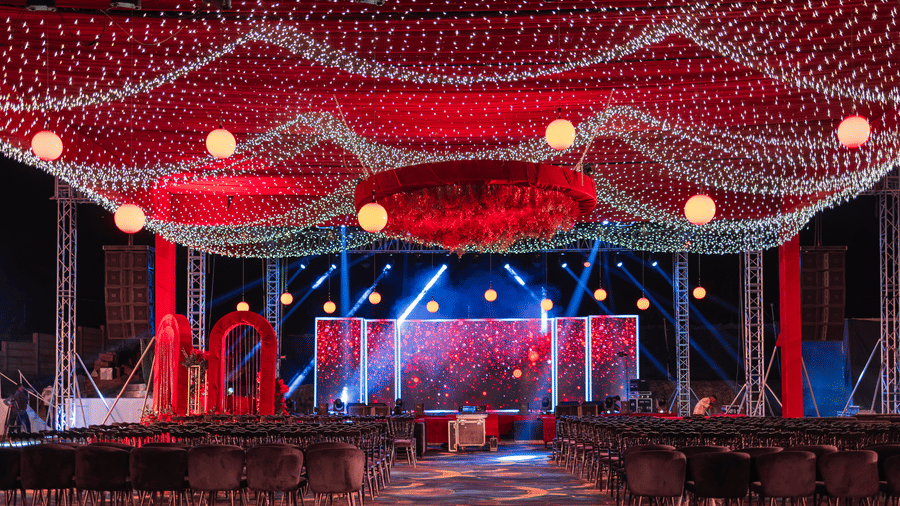 A wedding stage is decorated with red draping and lights indoors | The Qualia Resort Club & Brewery, Udaipur