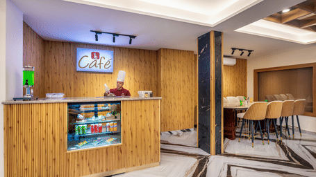 The reception area at Lords Inn Katra featuring a wooden counter, a glass display fridge, a television screen, and a seating section.