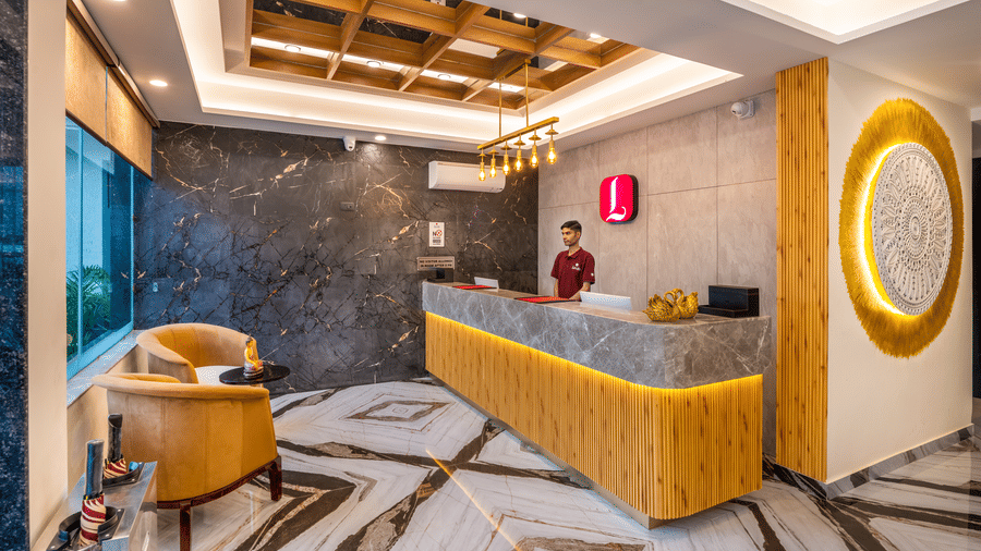 The main reception desk at Lords Inn Katra includes a long wooden counter, a textured dark feature wall, and a circular illuminated wall mirror.