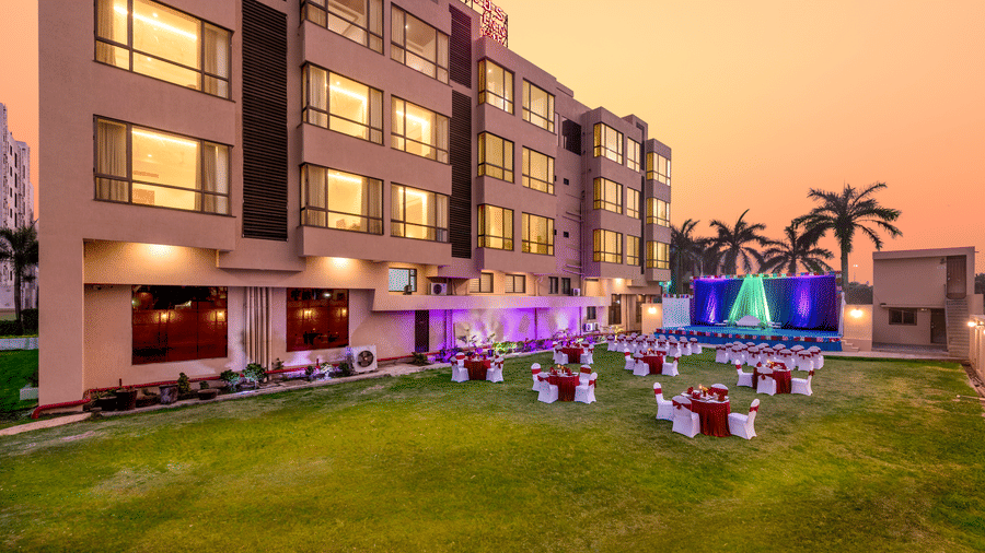 Evening view of the outdoor banquet lawn at Lords Inn Somnath, set up with round dining tables draped in white and red covers, decorative lighting along the building, and a vibrant stage with green and blue drapes.