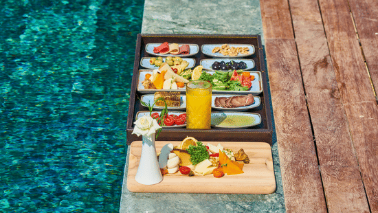 Floating breakfast tray with fruit, nuts, olives, juice and charcuterie set on pool edge with wooden side board.