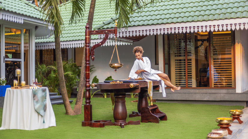 A woman in bathrobe getting on a spa bed which is placed in a lawn of Heritage Village Resort and Spa, Goa. The lawn also has other spa products. 