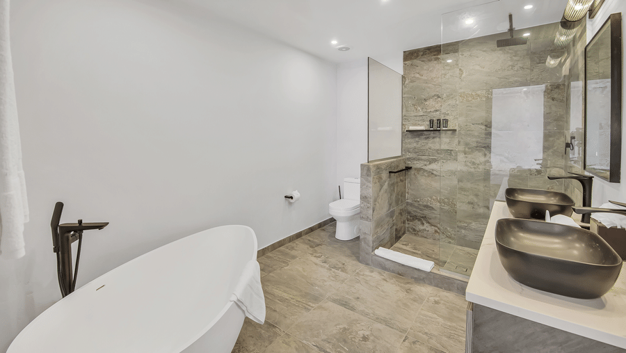 A bathroom at Tamarind Hills Resort and Villas, Antigua, with a bathtub with a faucet, two sinks with a mirror, a shower area and a toilet.
