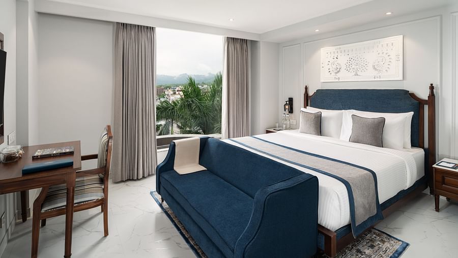 A room with a large bed with a blue bed runner, placed in front of a work desk, and a window offering a scenic view - MJ Sarovar Portico, Haridwar