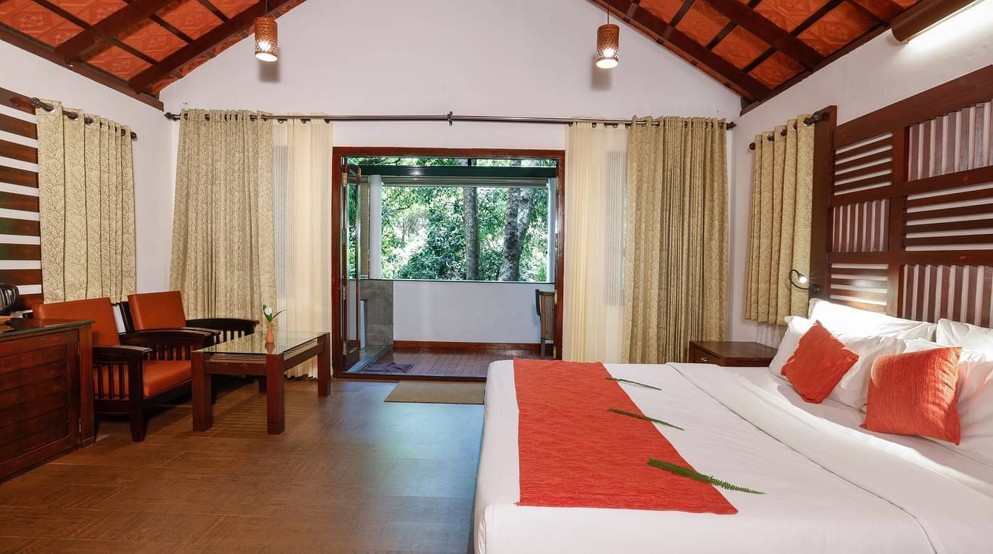 A side view of a room that has a bed, two chairs, and a balcony with forest views | Vythiri Resort, Wayanad