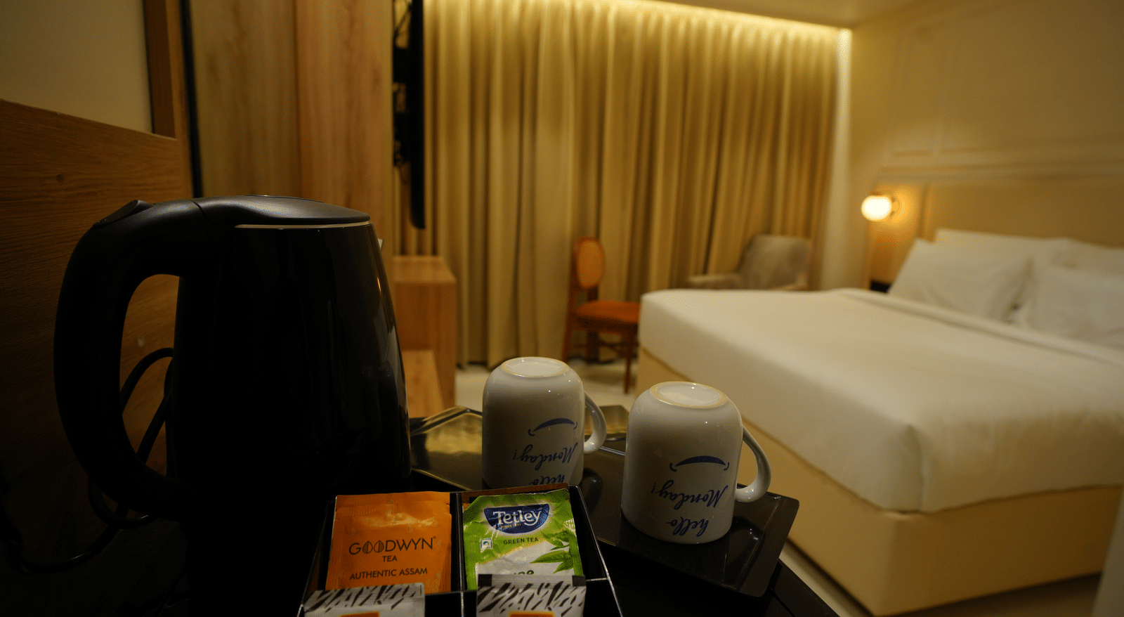  Kettle and mugs for room amenities at Monday Premium, Madhapur, with the double bed and gold-draped window blurred in the background.