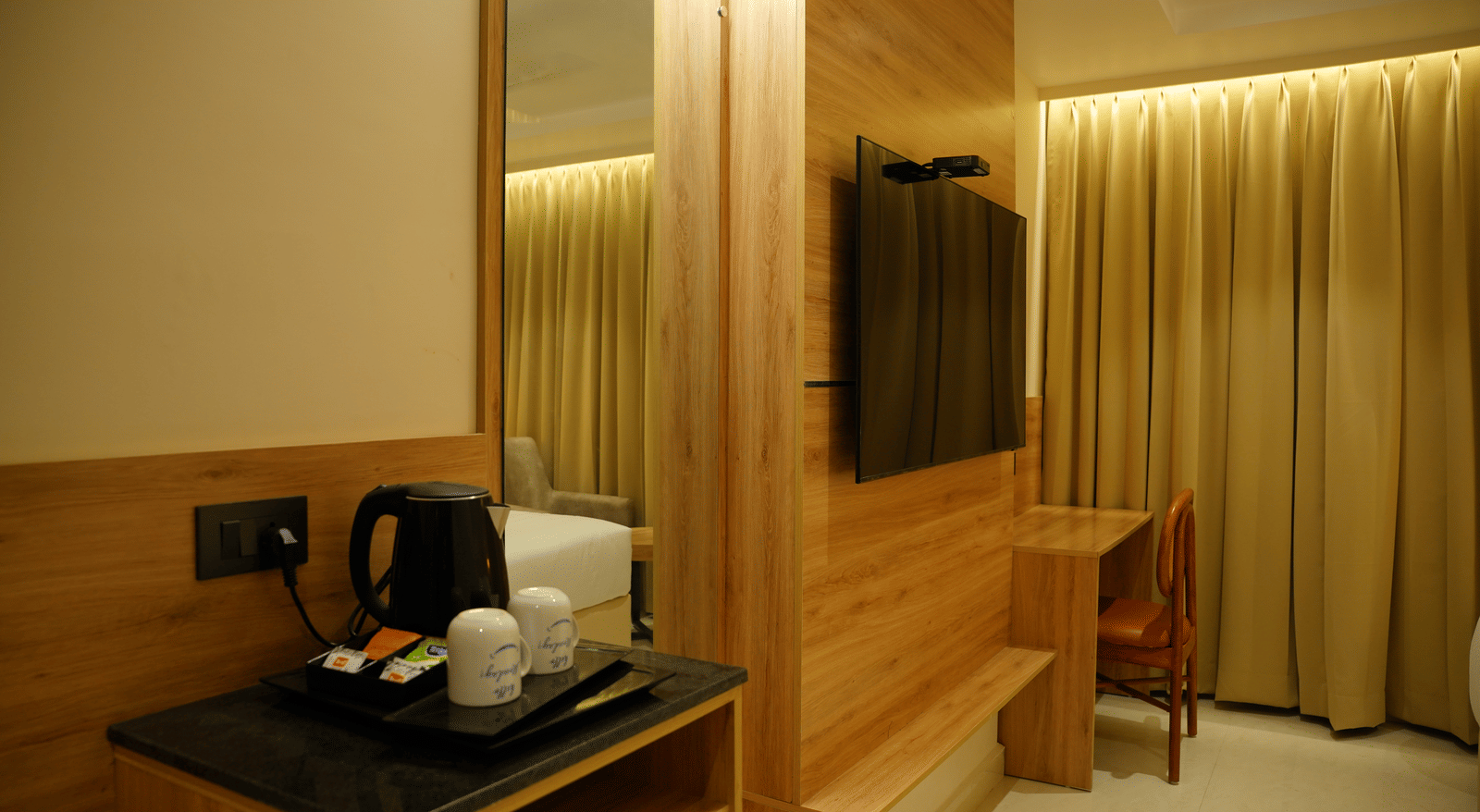  Modern guest room setup at Monday Premium, Madhapur, showing a TV on a wood-panelled wall and tea-making facilities nearby.