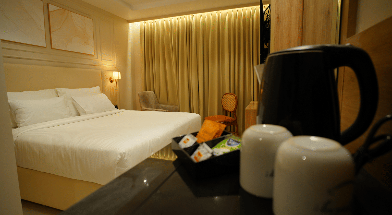  Room amenities at Monday Premium, Madhapur, featuring a kettle and two mugs out of focus, with the king-sized bed visible beyond.