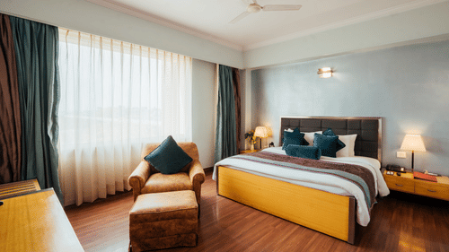 A bedroom at The Piccadily Lucknow featuring a bed, a sofa chair, bedside table with lamps on either side, and a large window with curtains.