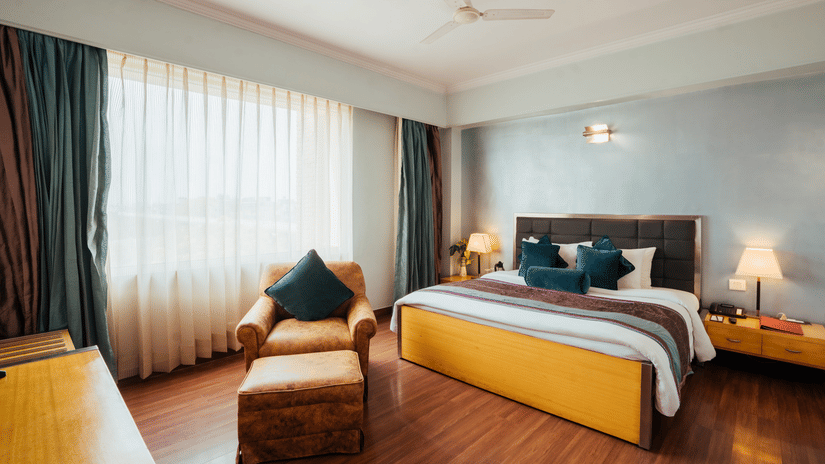 A room at The Piccadily Lucknow hotel featuring a double size bed, a sofa chair, bedside table with lamps on either side, and a large window with curtains.