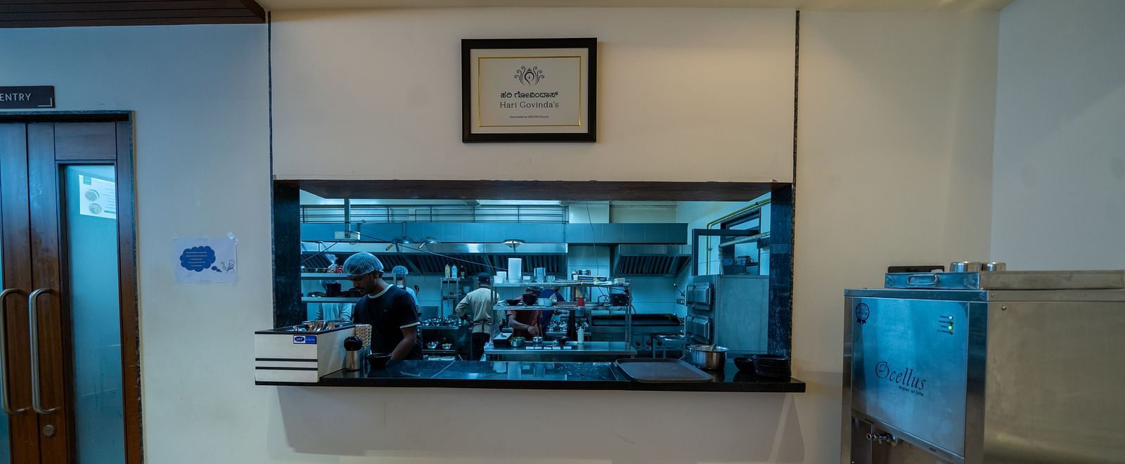 A rectangular glass on the wall at Hari Govinda's offering a view of the other side, with a photo frame above it and a metal water purifier beside it