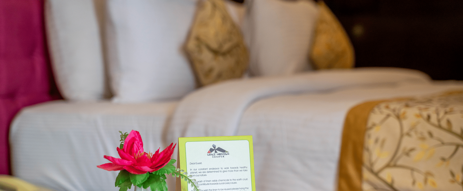 A neatly made hotel bed with white linens, decorative pillows, and a bedside table holding a flower vase and a service card - Hotel Udai Median, Udaipur.