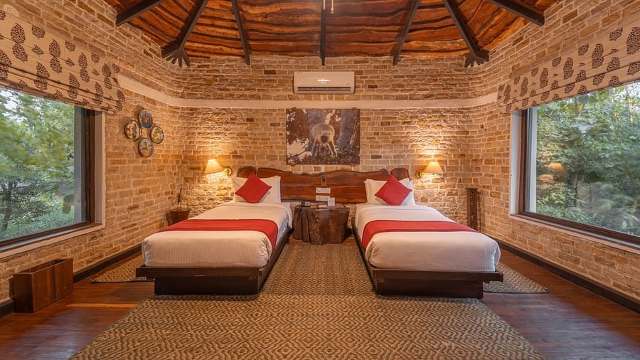 A bedroom at Tendu Leaf Jungle Resort featuring twin beds, large window on wither side , a bedside table, and a ceiling light in a wooden furnished room.