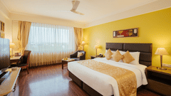 A spacious bedroom with a yellow feature wall, a bed with a golden runner, writing desk, armchair, and sheer window drapes at The Piccadilly Lucknow.