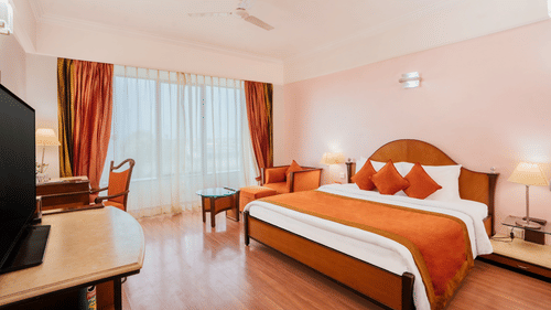 A warm-toned bedroom with a wooden bed, matching runner and cushions, work table, and a large window at The Piccadilly Lucknow.