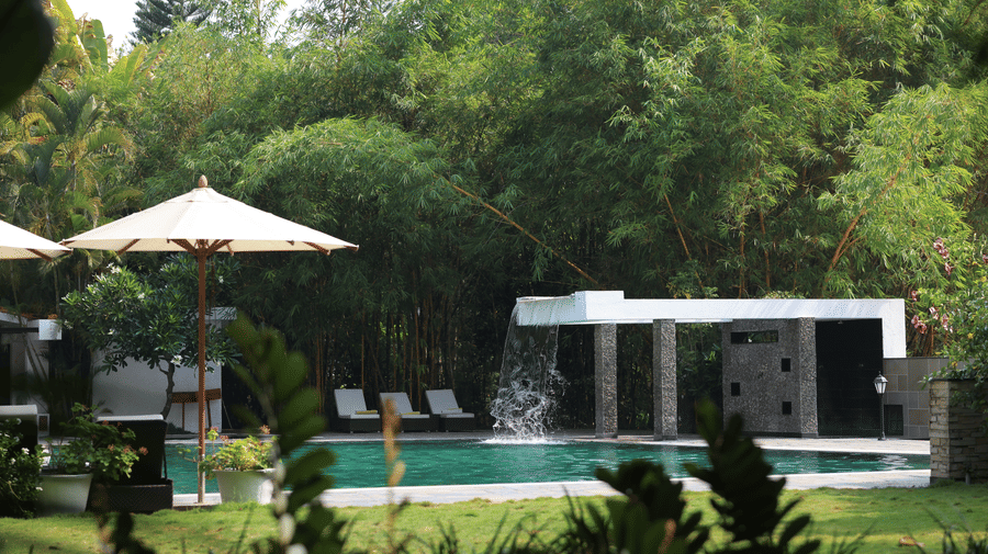 Poolside area at Amanvana Spa Resort - A Luxury Resort in Coorg featuring loungers, umbrellas, bamboo trees, and a waterfall feature.