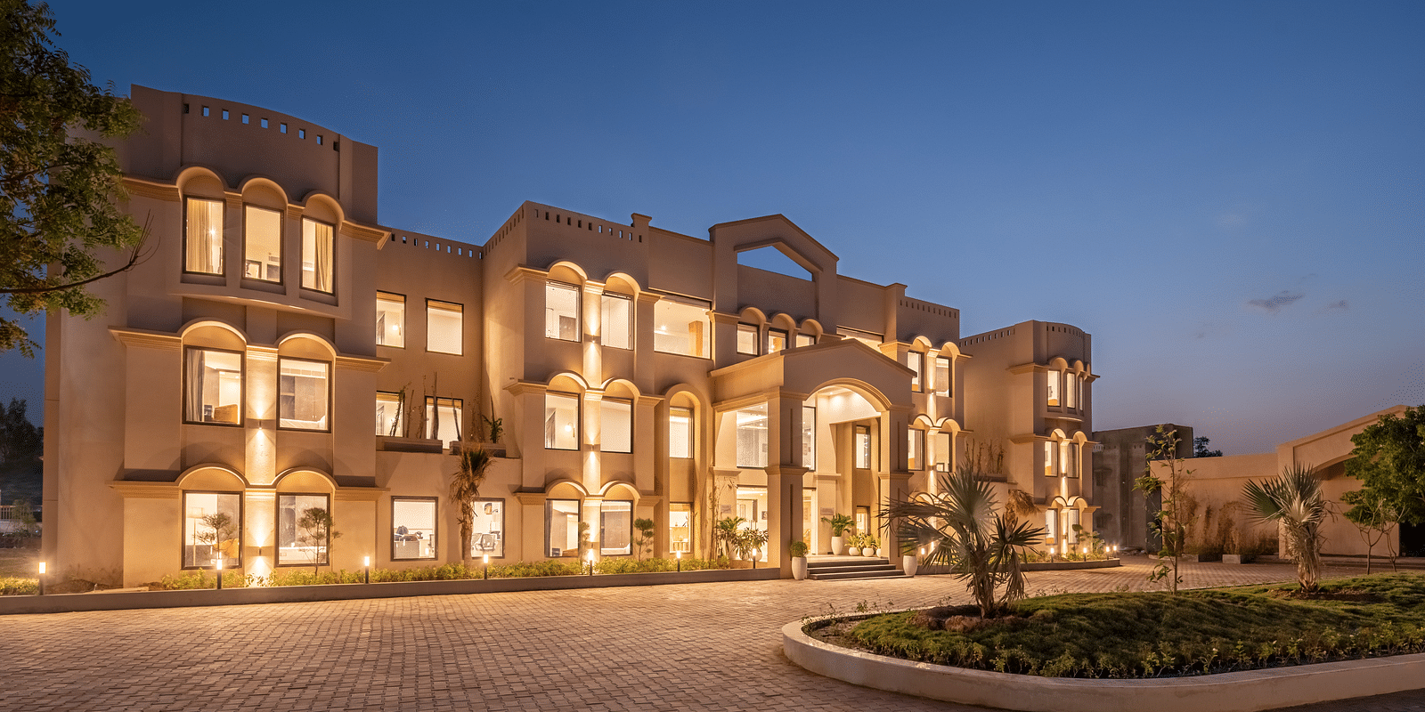 An image of a well lit facade with a huge concrete lawn and a few trees - Brij Eternity, Vrindavan