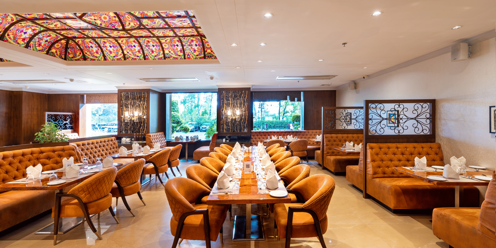 Restaurant dining hall featuring orange booths, wooden tables set with orange cloths, and a decorative ceiling panel at Hotel Hukam's Lalit Mahal.